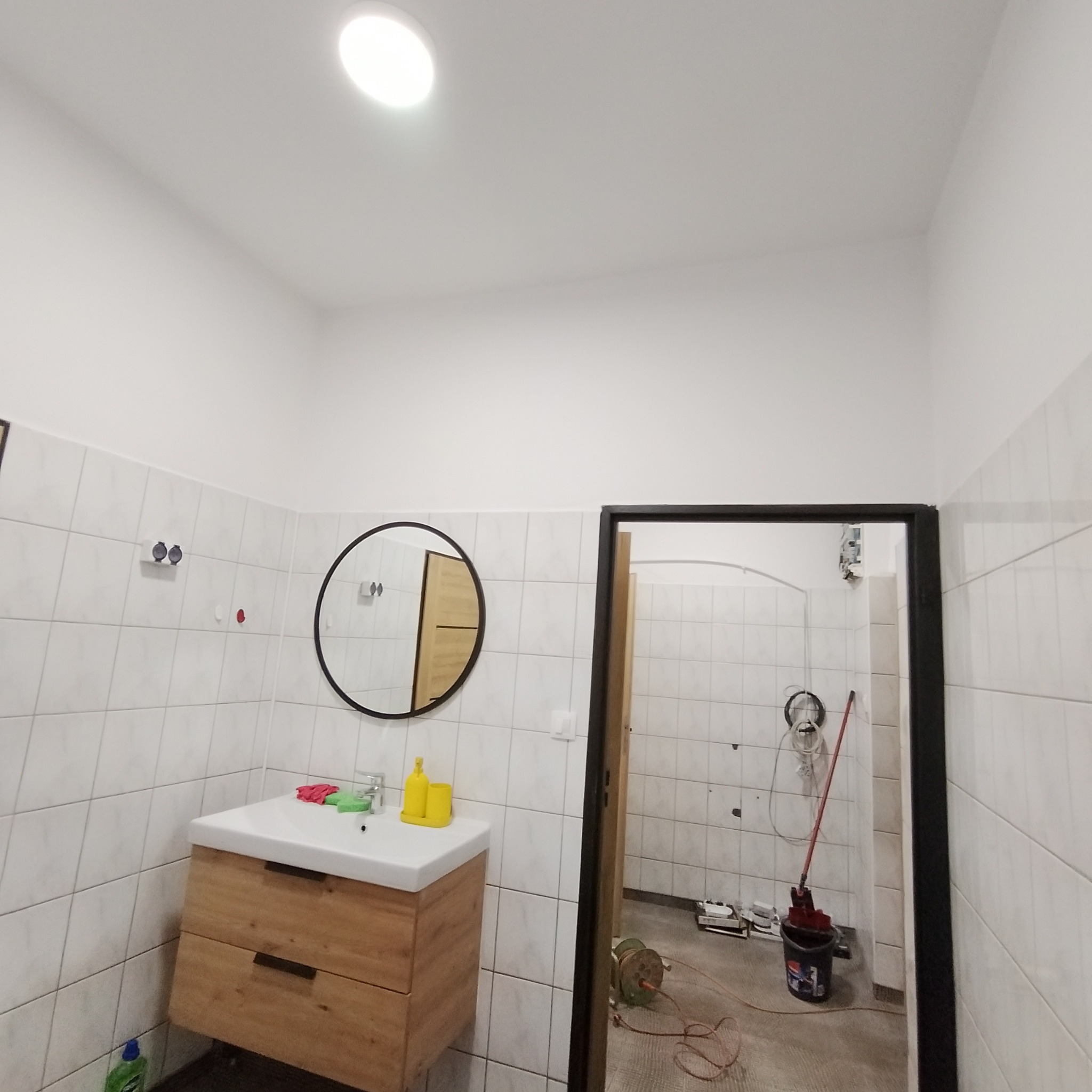Łazienka w trakcie remontu: białe płytki, drewniana szafka z umywalką, okrągłe lustro, widoczne narzędzia i wiadro z mopem w przejściu do innego pomieszczenia.