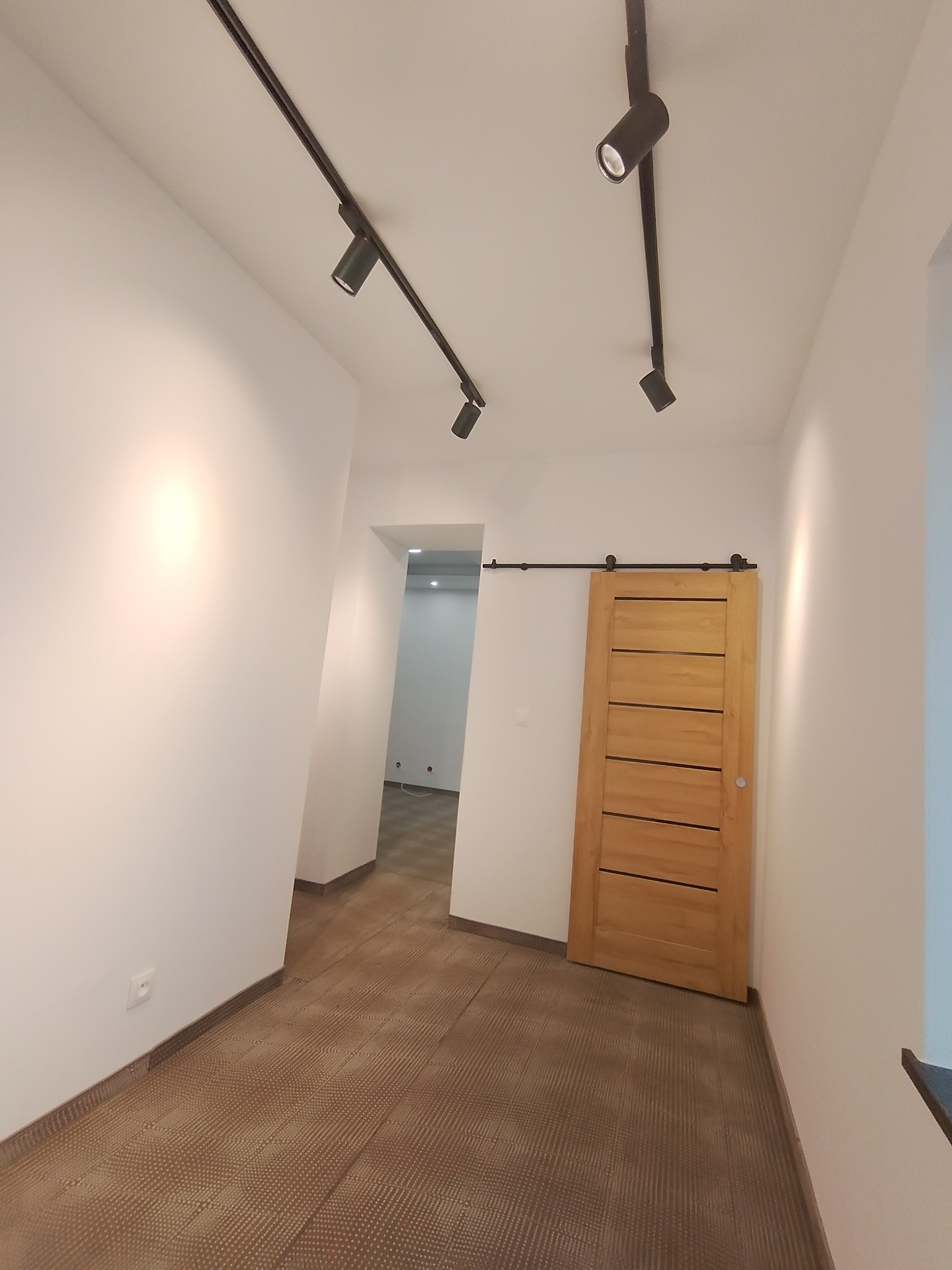 Jasne wnętrze z panelami podłogowymi w odcieniach brązu, przesuwnymi drewnianymi drzwiami i czarnymi reflektorami sufitowymi na szynoprzewodzie.