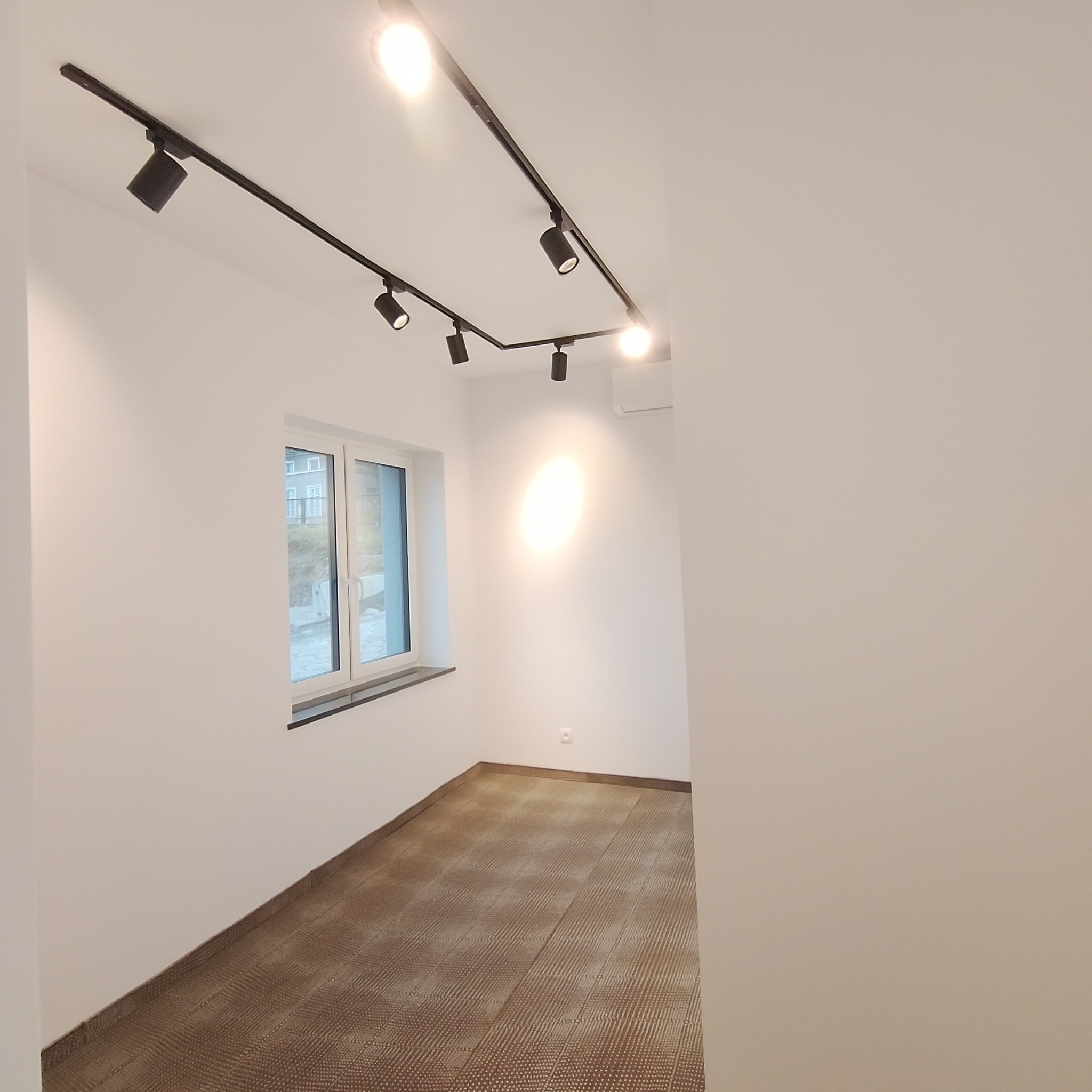 Jasne, puste pomieszczenie z nową podłogą imitującą drewno, oświetleniem szynowym na suficie i klimatyzatorem, widok na okno z zewnątrz.