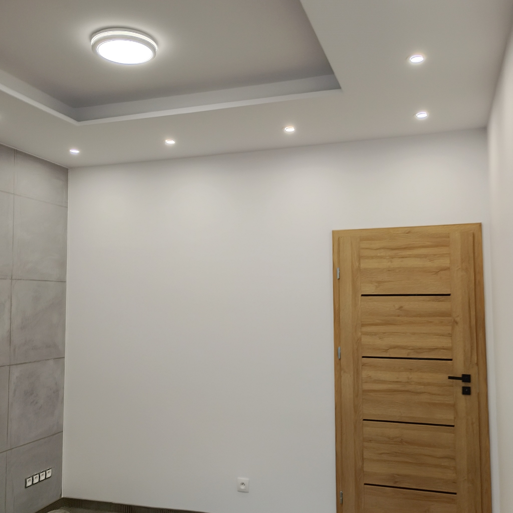Wnętrze pokoju po remoncie, z szarą ścianą z płytek imitujących beton, gładką białą ścianą, drewnianymi drzwiami z czarną klamką i nowoczesnym oświetleniem sufitowym.