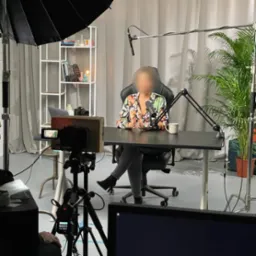 Kobieta w kwiecistej bluzce siedzi za biurkiem z mikrofonem w studio nagraniowym z oświetleniem, półką z rekwizytami i rośliną doniczkową w tle, nagrywana przez kamerę na statywie.