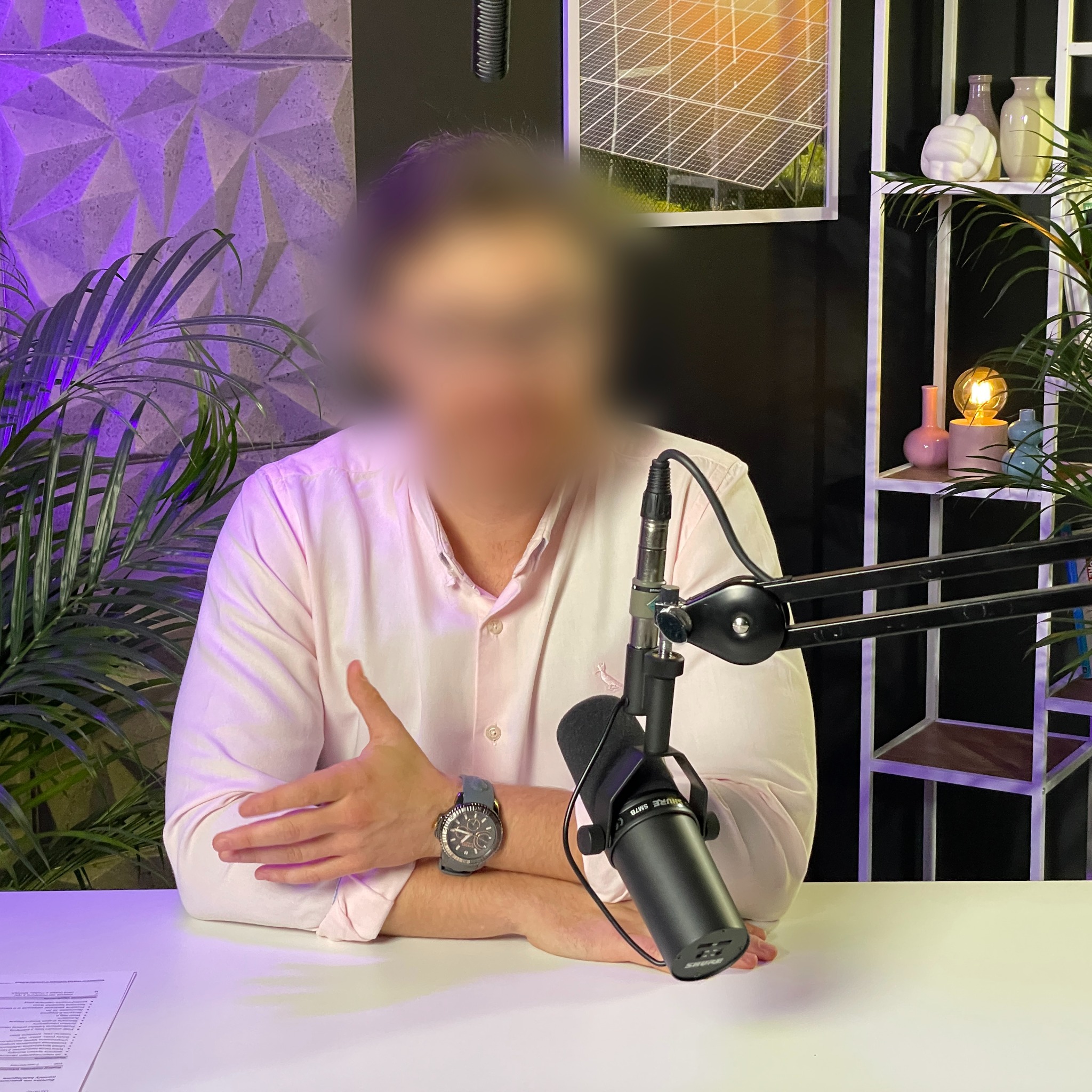 Osoba z zamazaną twarzą siedzi przy biurku z mikrofonem Shure SM7B, w tle roślina, geometryczna ściana podświetlona na fioletowo oraz półka z dekoracjami i obrazem paneli słonecznych.