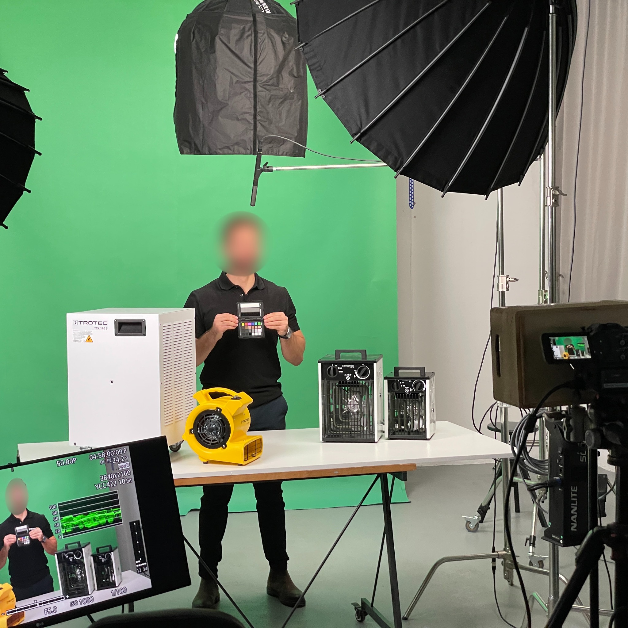 Sesja zdjęciowa z zielonym tłem: mężczyzna trzymający kartę kolorów, obok biały osuszacz powietrza Trotec TTK 140 S, żółta dmuchawa i dwa grzejniki elektryczne, oświetlenie studyjne i kamera...