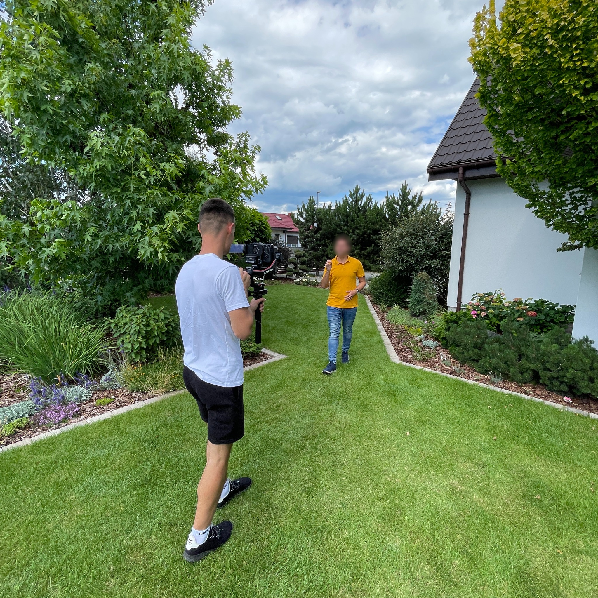 Operator kamery nagrywa osobę w żółtej koszulce na zadbanym trawniku przed domem z brązowym dachem.