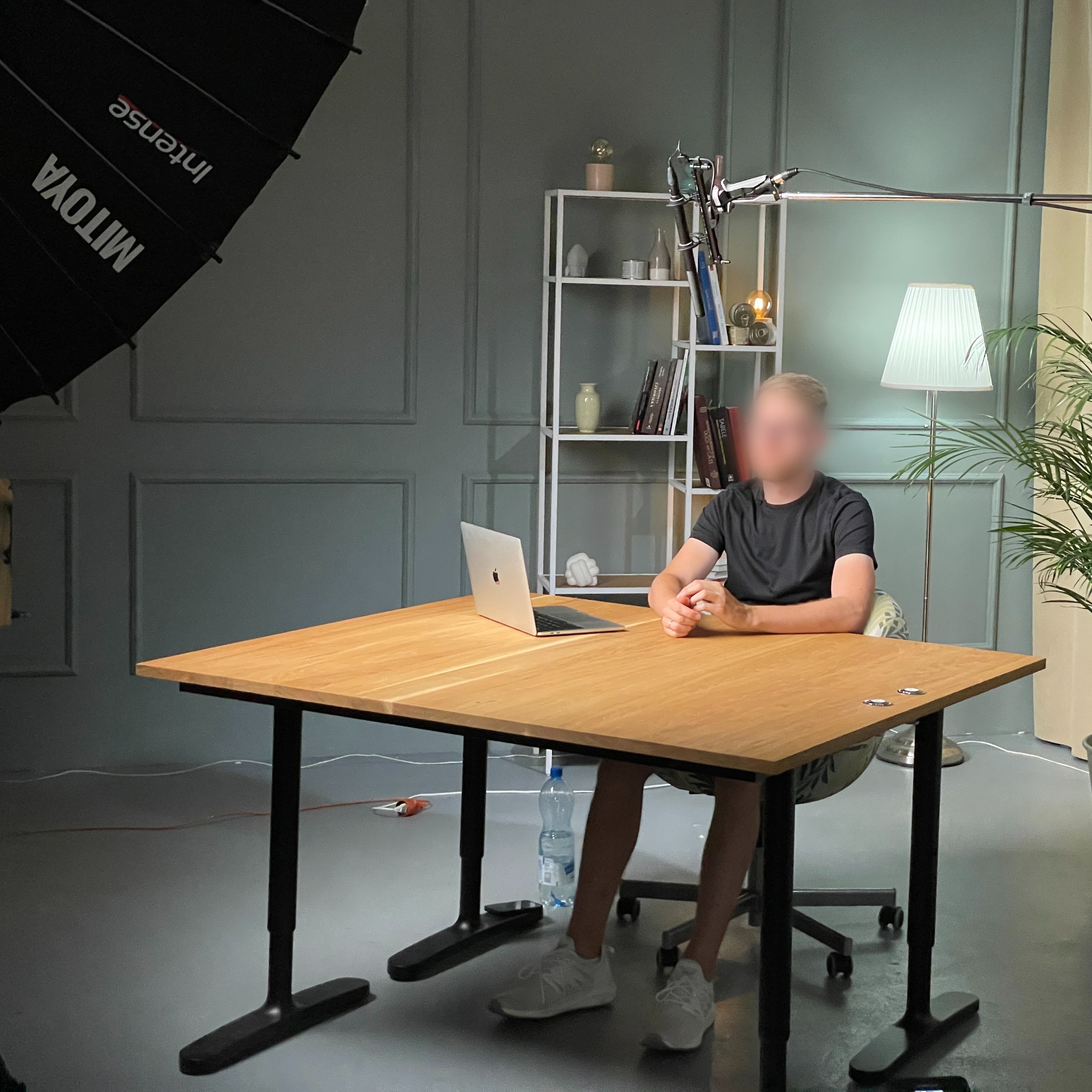 Osoba siedzi przy biurku z laptopem w studio oświetlonym lampą studyjną Mitoya Intense, z półką z książkami i lampą podłogową w tle.