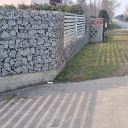 Paweł Fronc - Nowoczesne ogrodzenie gabionowe z metalową bramą i elementami drewnianymi, otoczone zielenią. Widok na ulicę i domy w tle, w słoneczny dzień.