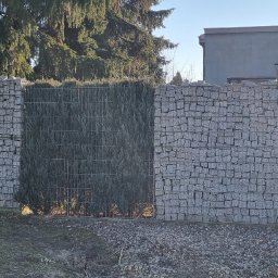 Paweł Fronc - Gabionowe ogrodzenie z siatki wypełnione kamieniem, przeplatane żywopłotem z tuj, na tle domu i drzew, z żółtą skrzynką gazową po lewej stronie.