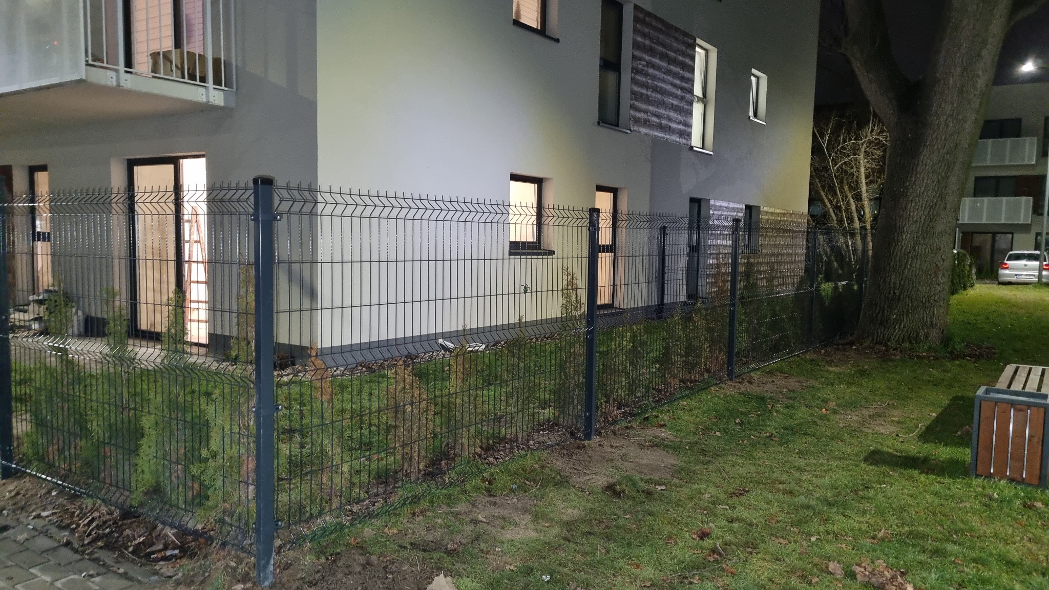 Nowoczesne ogrodzenie panelowe w kolorze grafitowym okalające budynek mieszkalny z małymi tujami posadzonymi wzdłuż niego, widoczne oświetlenie zewnętrzne i ławka parkowa.