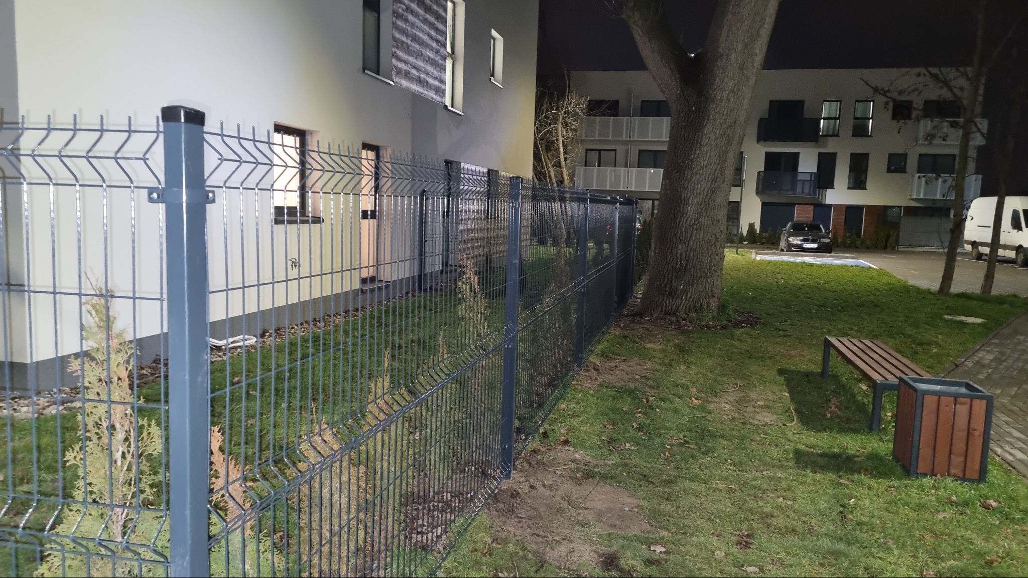 Szare, panelowe ogrodzenie z siatki zgrzewanej, oddzielające budynek mieszkalny od trawnika z drzewem i ławką, widoczne małe tuje posadzone wzdłuż ogrodzenia, nocne oświetlenie.
