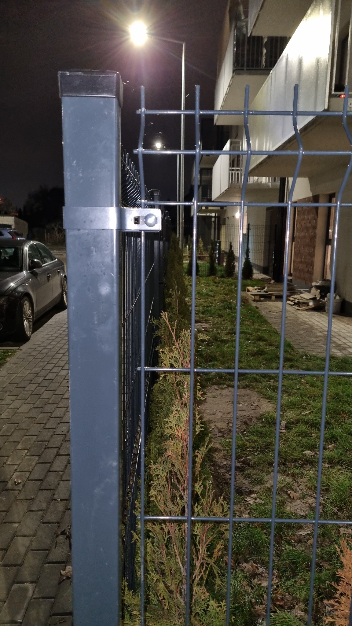 Szary metalowy słupek ogrodzeniowy z siatką na tle bloku mieszkalnego i chodnika z kostki brukowej, oświetlony uliczną lampą w nocy.