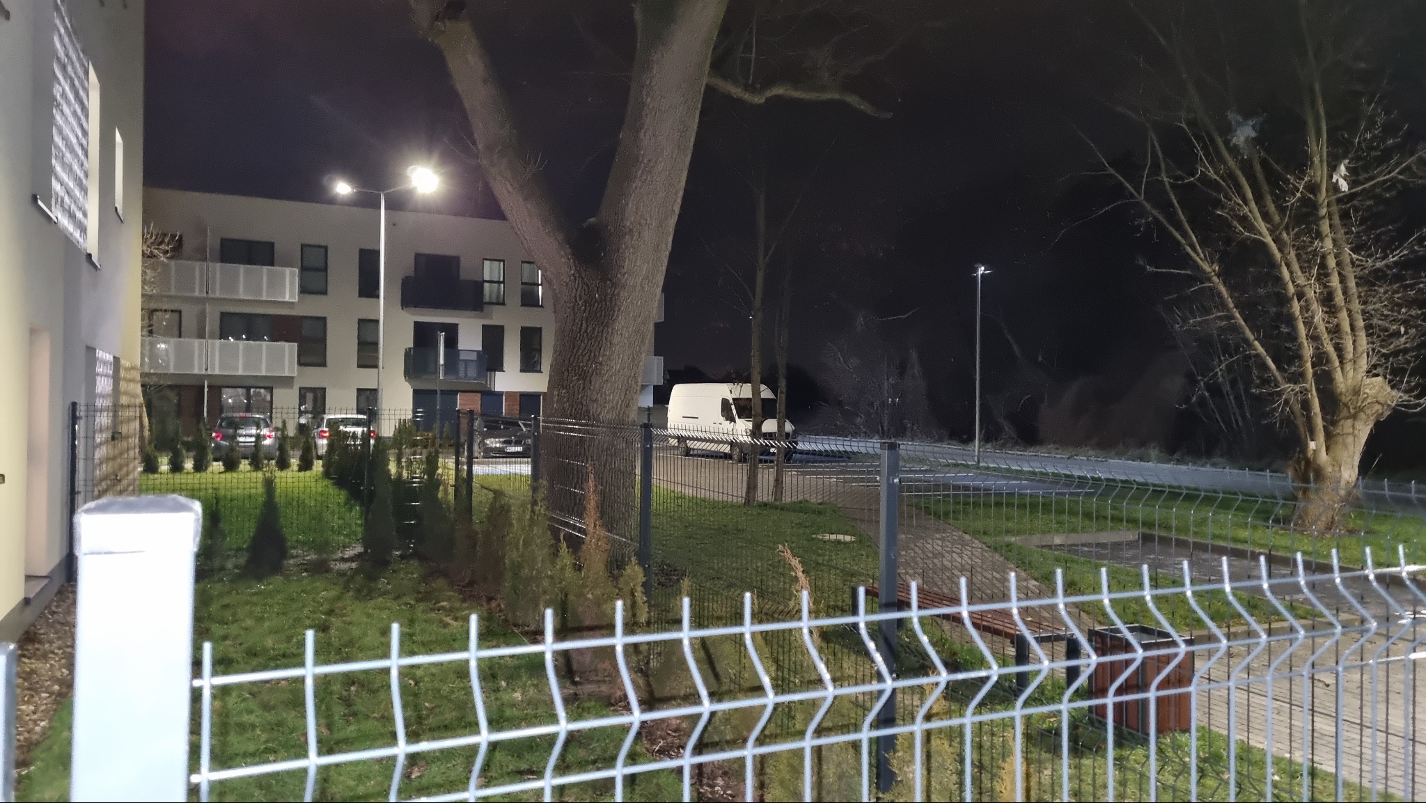 Nocne ujęcie metalowego ogrodzenia panelowego na tle oświetlonego bloku mieszkalnego, z widocznym samochodem dostawczym i drzewami.