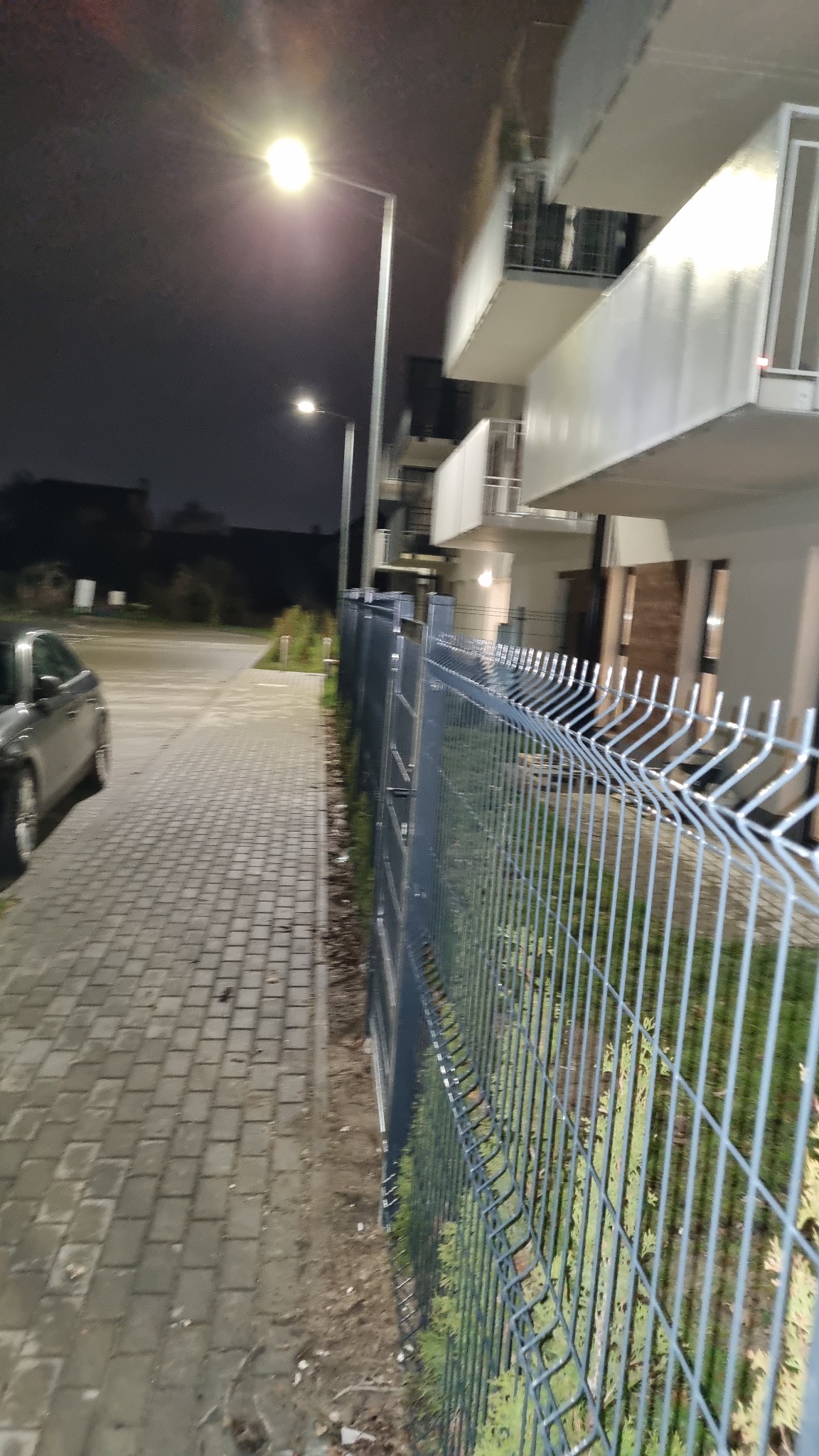 Szara, metalowa siatka ogrodzeniowa z widocznymi pionowymi prętami na tle chodnika z kostki i budynku mieszkalnego oświetlonego latarnią uliczną nocą.