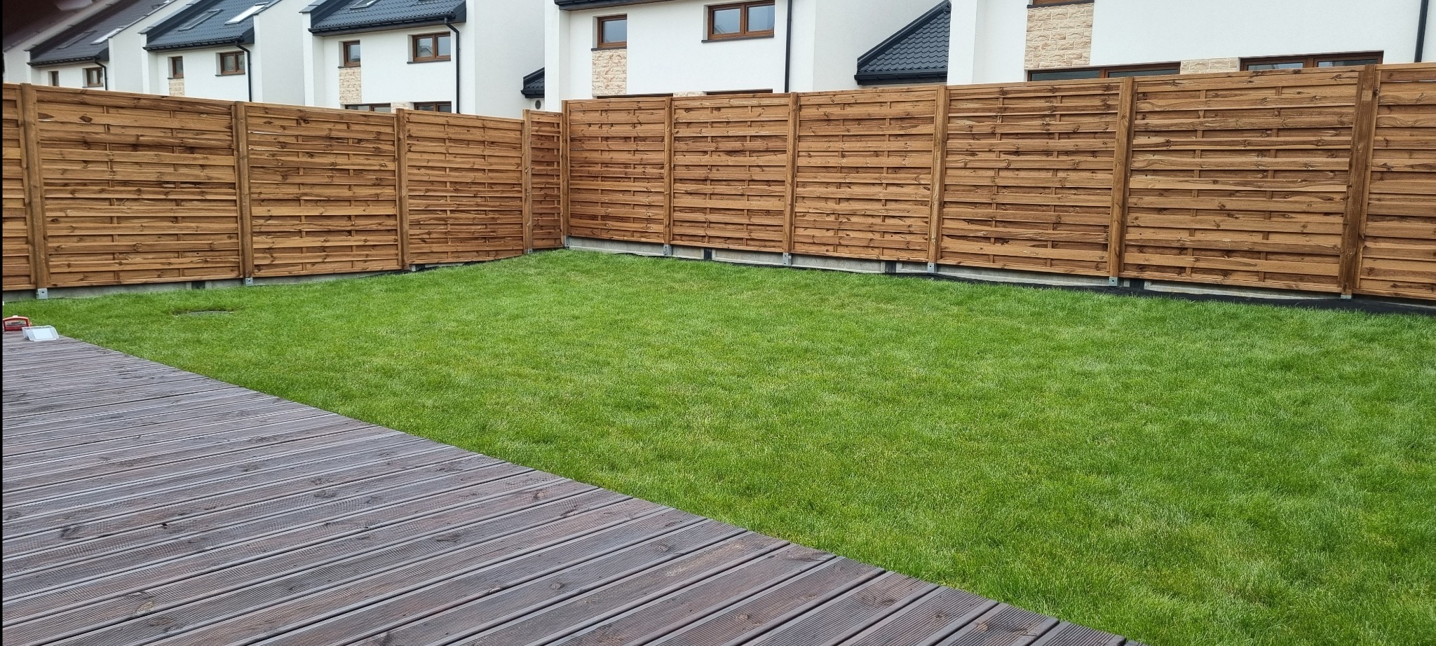 Nowoczesne, poziome ogrodzenie drewniane w kolorze brązowym, oddzielające trawnik od zabudowy szeregowej, widoczne z perspektywy drewnianego tarasu.