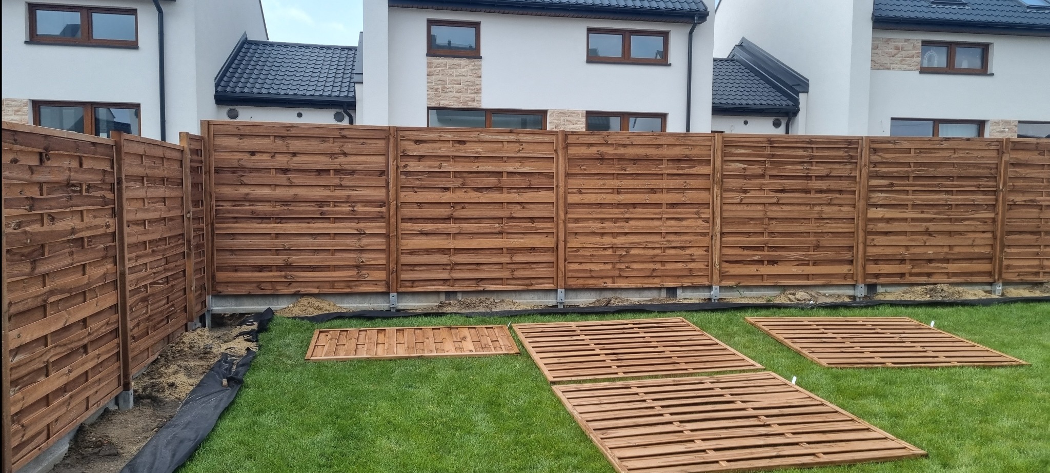 Montaż nowoczesnego, horyzontalnego ogrodzenia panelowego z drewna w kolorze brąz. Na trawie leżą przygotowane panele. W tle domy.