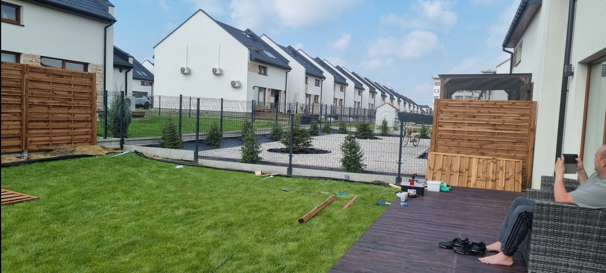 Widok na posesję z drewnianym ogrodzeniem panelowym i metalową siatką. W tle szereg identycznych domów, na pierwszym planie trawnik i taras z osobą.