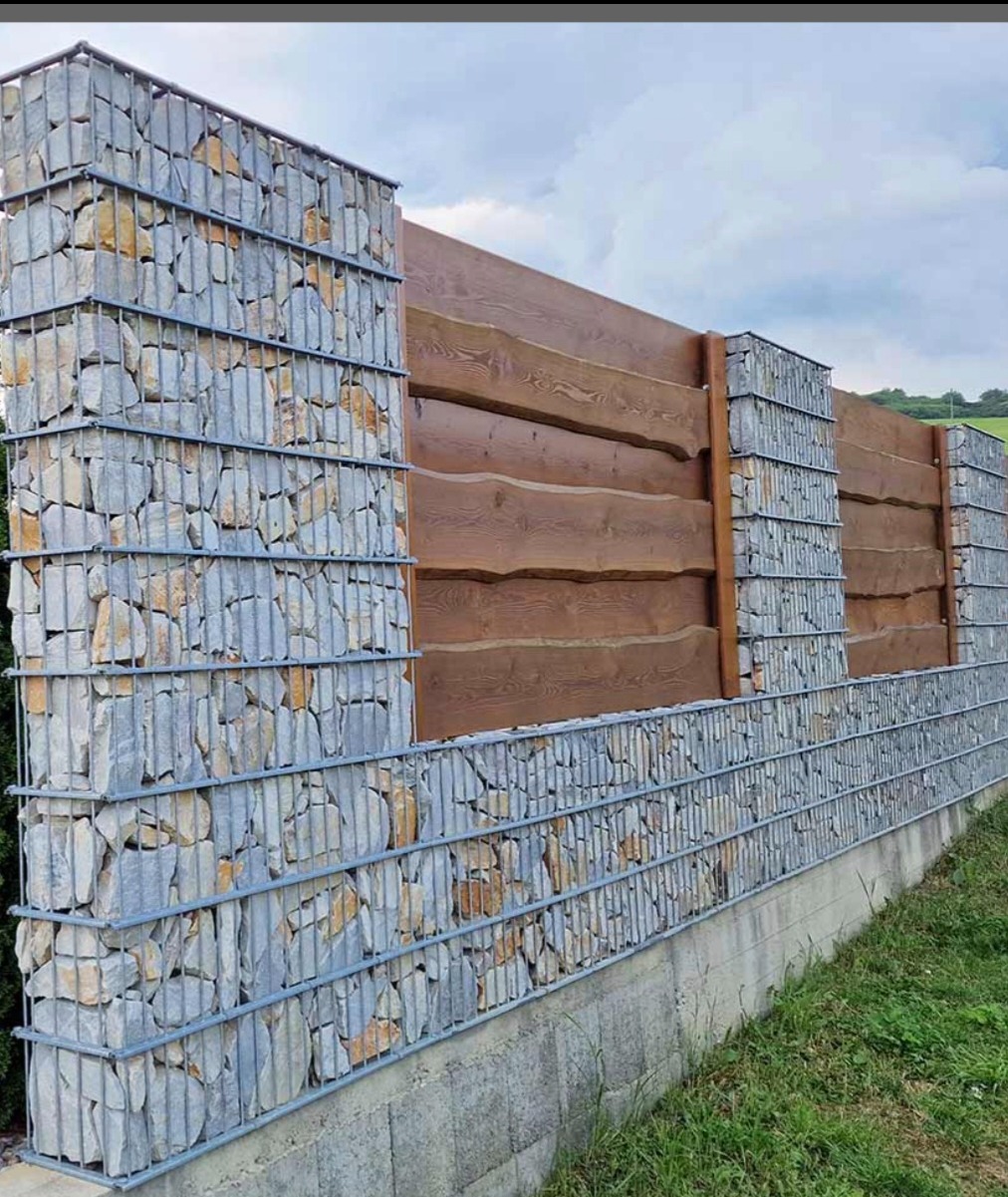 Nowoczesne ogrodzenie: gabiony wypełnione kamieniem połączone z drewnianymi panelami o nieregularnych krawędziach, na betonowej podmurówce, w tle zielony krajobraz.