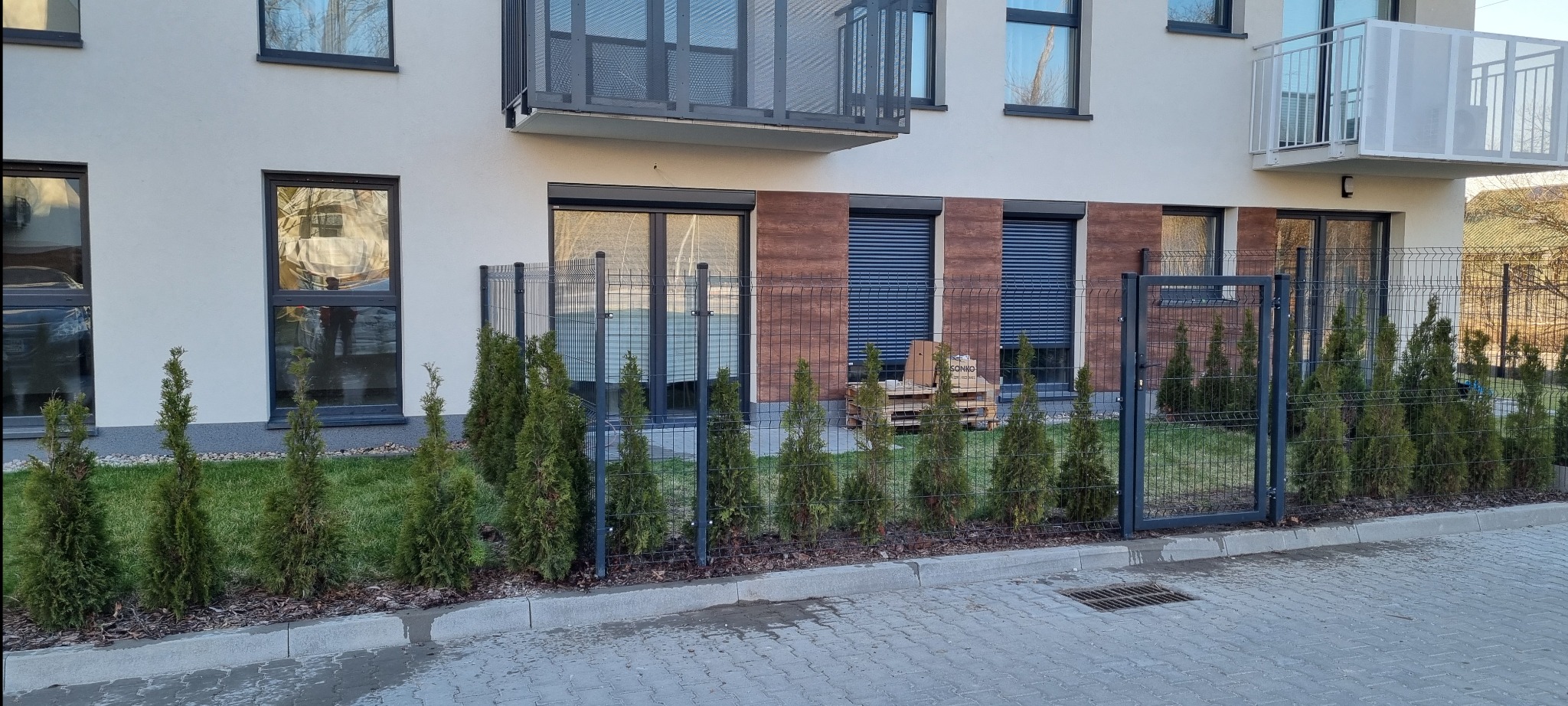 Nowoczesne ogrodzenie panelowe w kolorze grafitowym, oddzielające budynek od chodnika, z niskimi tujami wzdłuż. Widoczna brama wejściowa i paleta z materiałami budowlanymi.