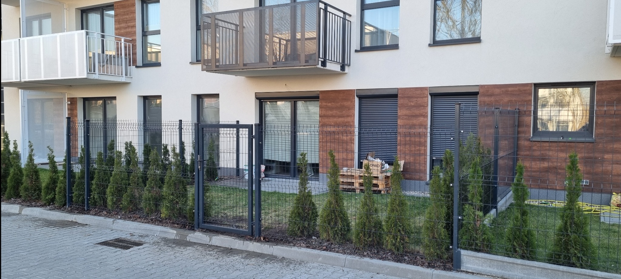 Nowoczesne ogrodzenie panelowe w kolorze grafitowym, otaczające budynek wielorodzinny, z niskimi tujami wzdłuż. Brama wejściowa zamyka posesję.