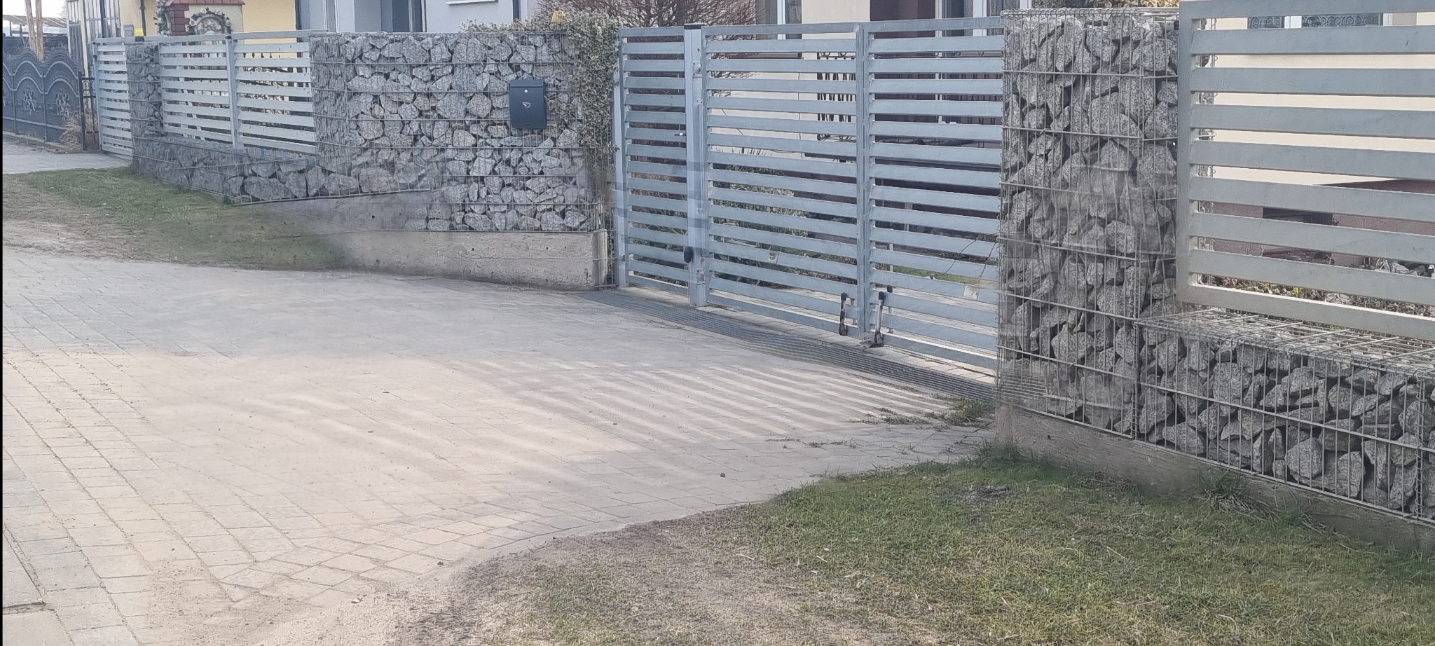 Nowoczesne ogrodzenie z metalowymi przęsłami i gabionami wypełnionymi kamieniami. Brama wjazdowa z automatyką. Skrzynka pocztowa na słupku.
