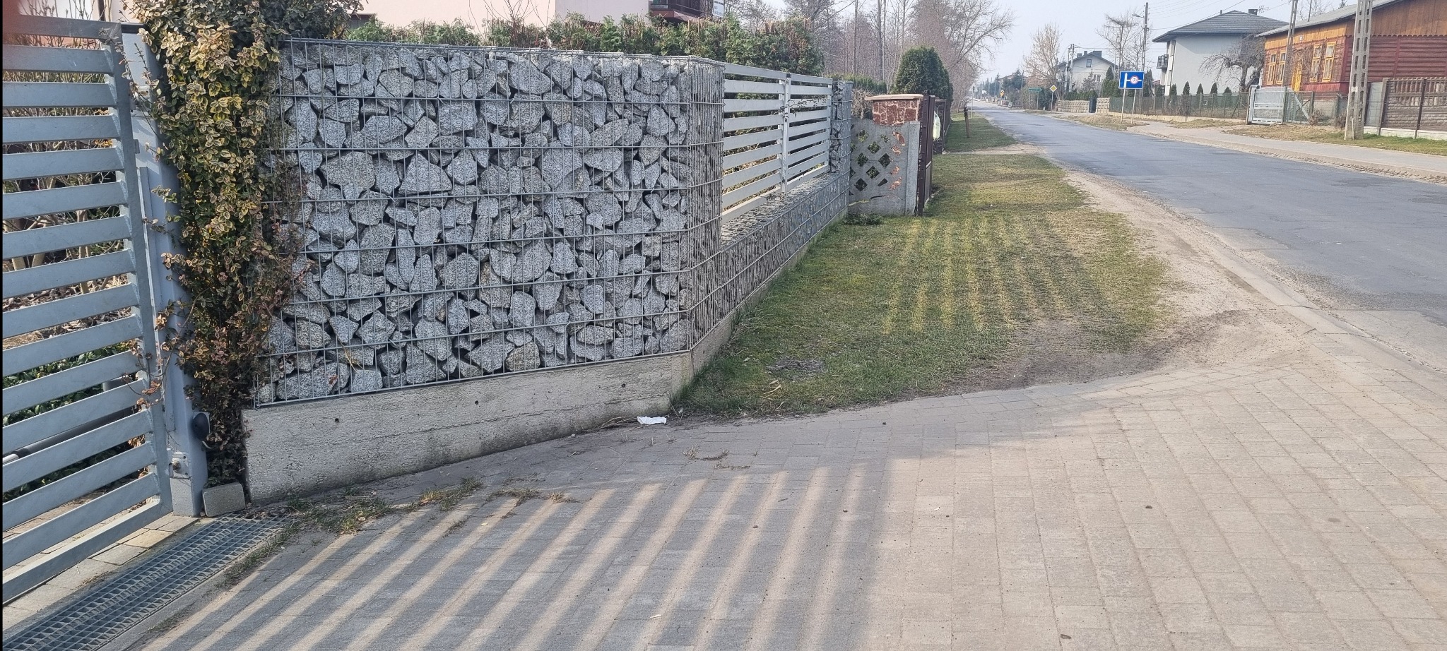 Nowoczesne ogrodzenie gabionowe z metalową bramą i elementami drewnianymi, otoczone zielenią. Widok na ulicę i domy w tle, w słoneczny dzień.