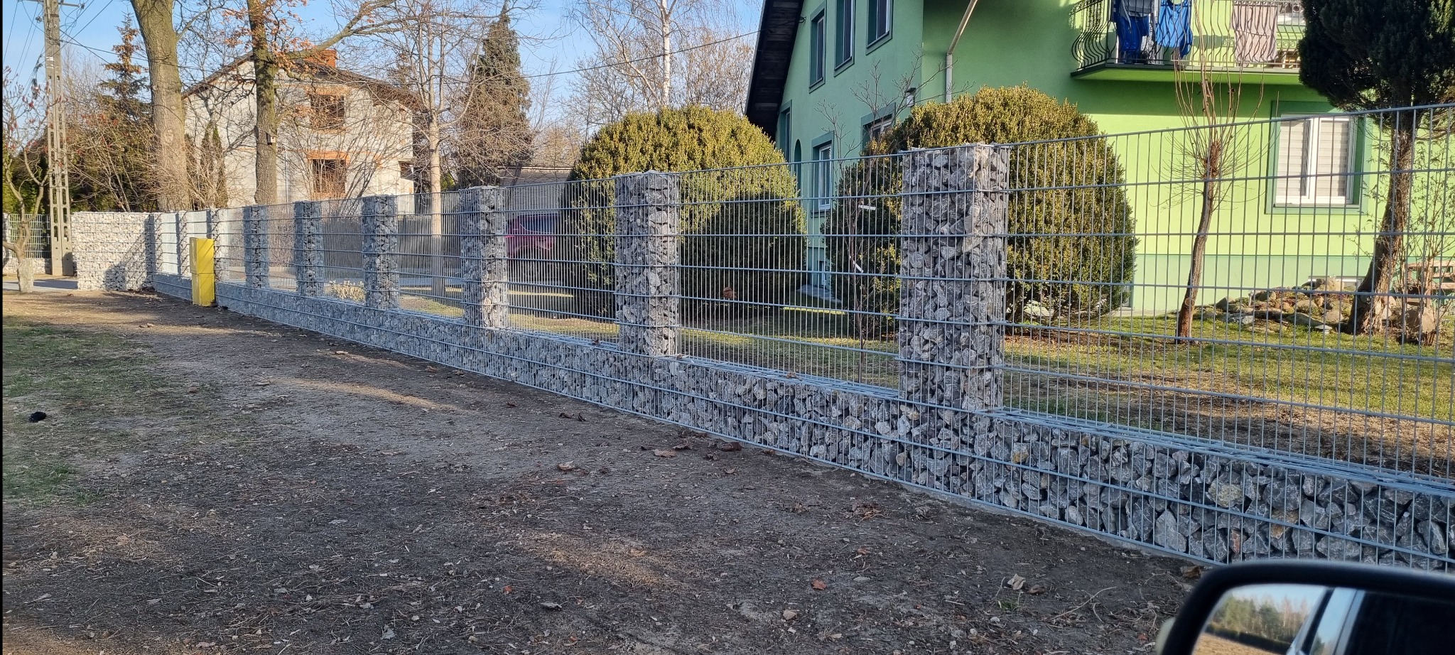 Gabionowe ogrodzenie z metalowej siatki wypełnionej kamieniami, oddzielające posesję. W tle domy i drzewa w słoneczny dzień. Widok z perspektywy ulicy.