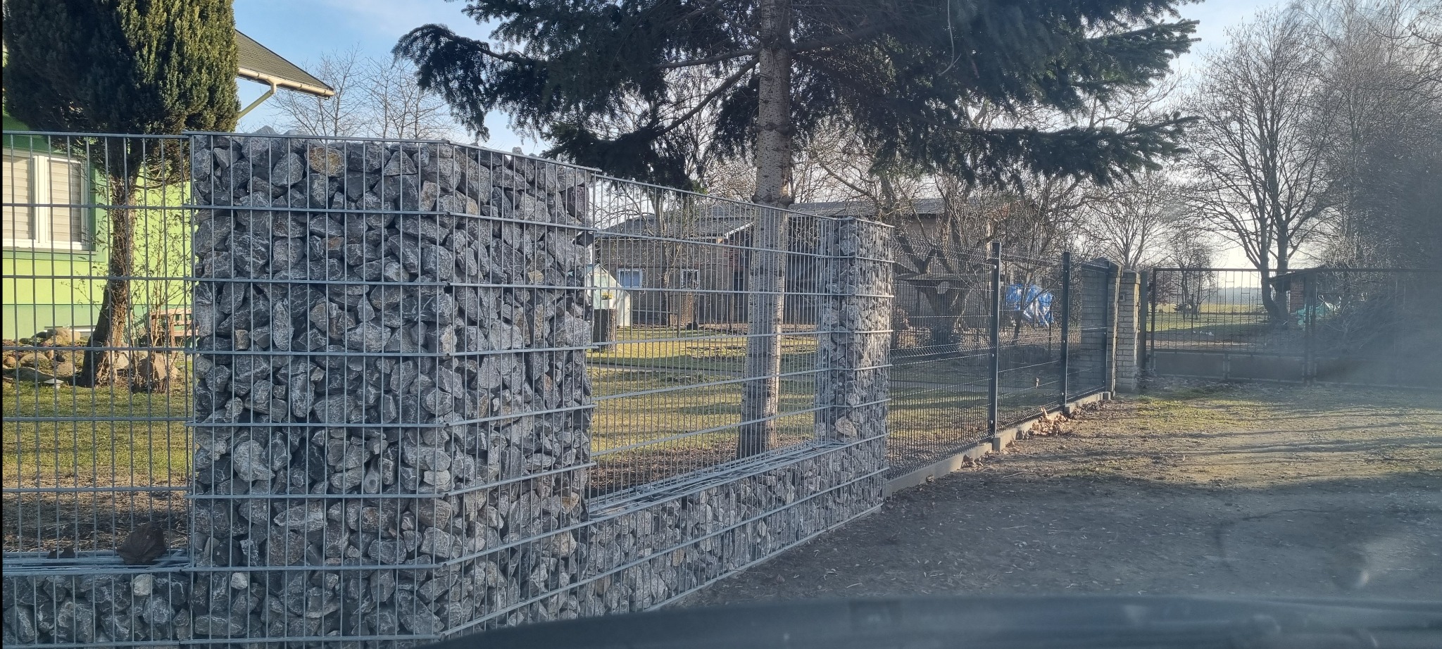 Gabionowy fragment ogrodzenia z siatki wypełniony kamieniami, tworzący solidną i estetyczną barierę w otoczeniu zieleni i zabudowań.