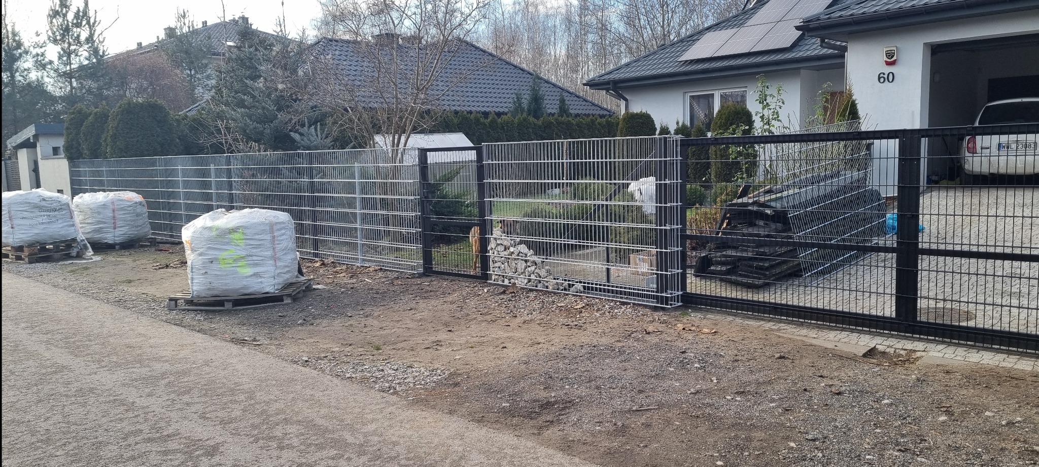 Nowoczesne ogrodzenie panelowe w kolorze szarym i czarnym, z widocznymi workami z materiałem budowlanym i posesją w tle, w słoneczny dzień.
