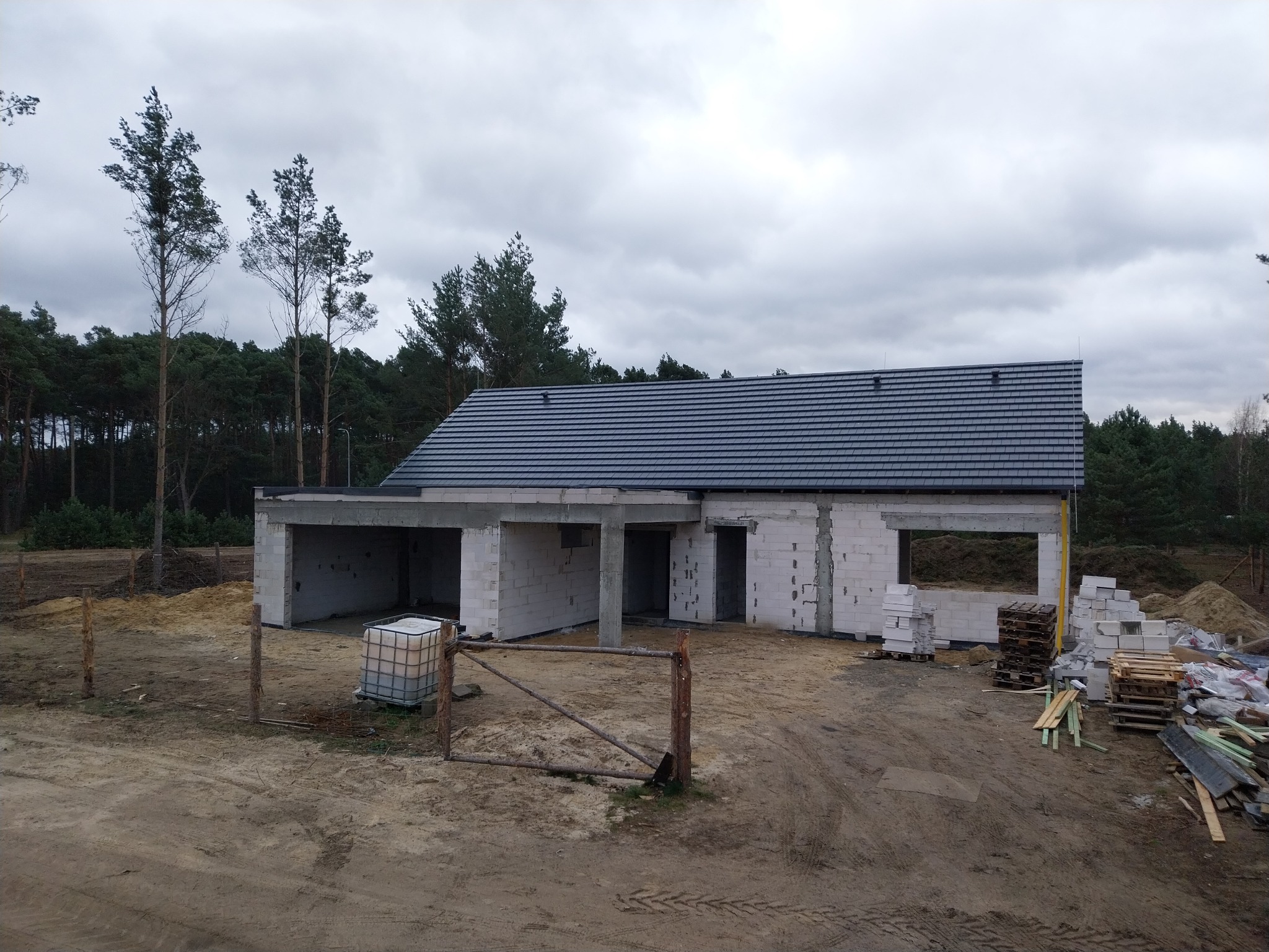 Surowy stan domu z betonu komórkowego z ciemną dachówką na tle pochmurnego nieba i lasu, widoczne palety z materiałami budowlanymi i tymczasowe ogrodzenie.