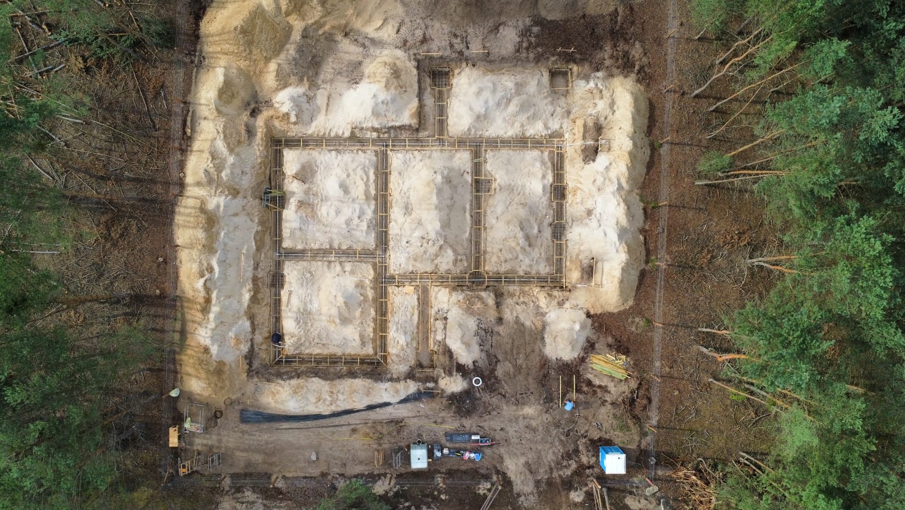 Widok z góry na wykop pod fundamenty z drewnianym szalunkiem, otoczony lasem iglastym i piaskiem.