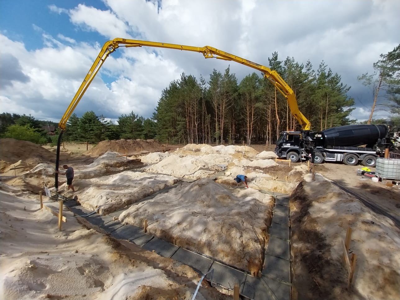 Nalewanie betonu do wykopanych fundamentów z użyciem pompy zamontowanej na ciężarówce z betonomieszarką, widoczni dwaj pracownicy w trakcie pracy.