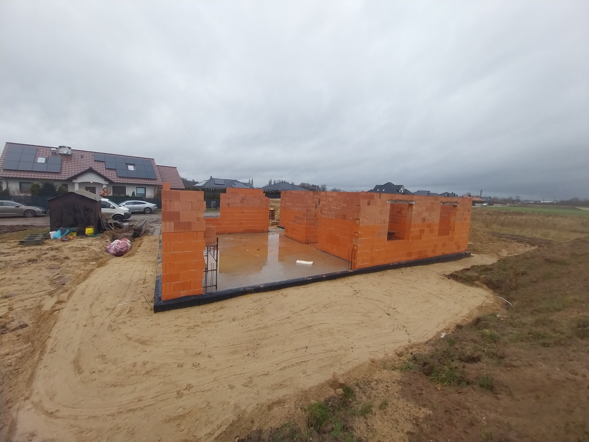 Wznoszenie ścian parteru z czerwonej cegły na fundamencie, widoczne zbrojenia, na zewnątrz piasek i budynki mieszkalne w tle, pochmurny dzień.