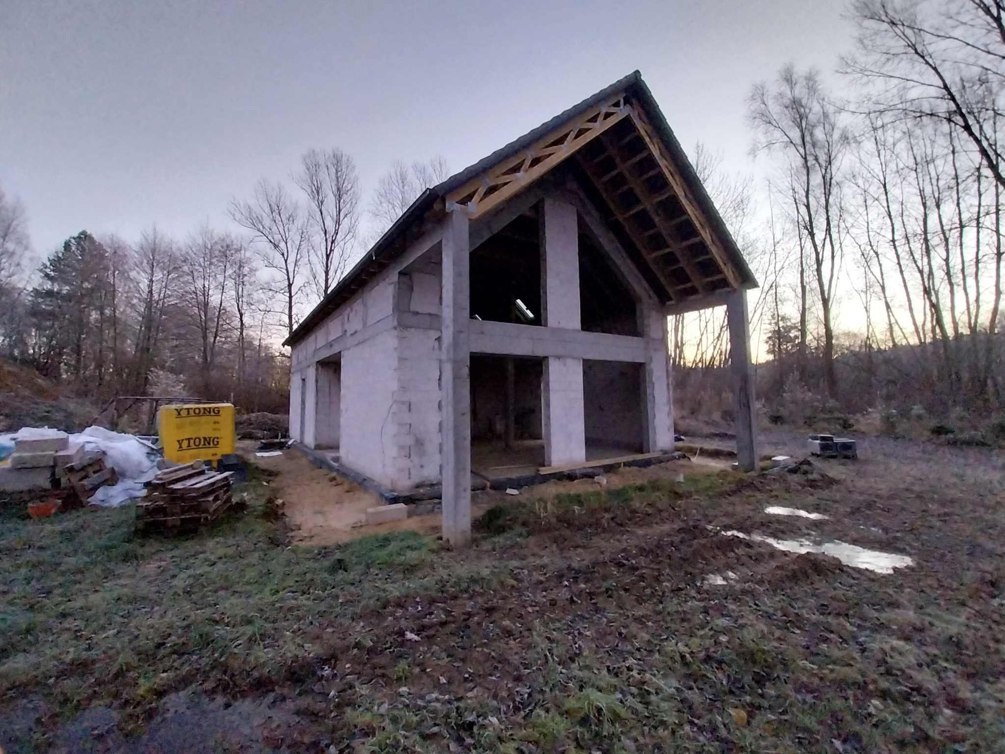 Surowy stan domu z betonu komórkowego z widoczną więźbą dachową, bez okien i drzwi, na tle jesiennego krajobrazu, widoczne palety z bloczkami Ytong.