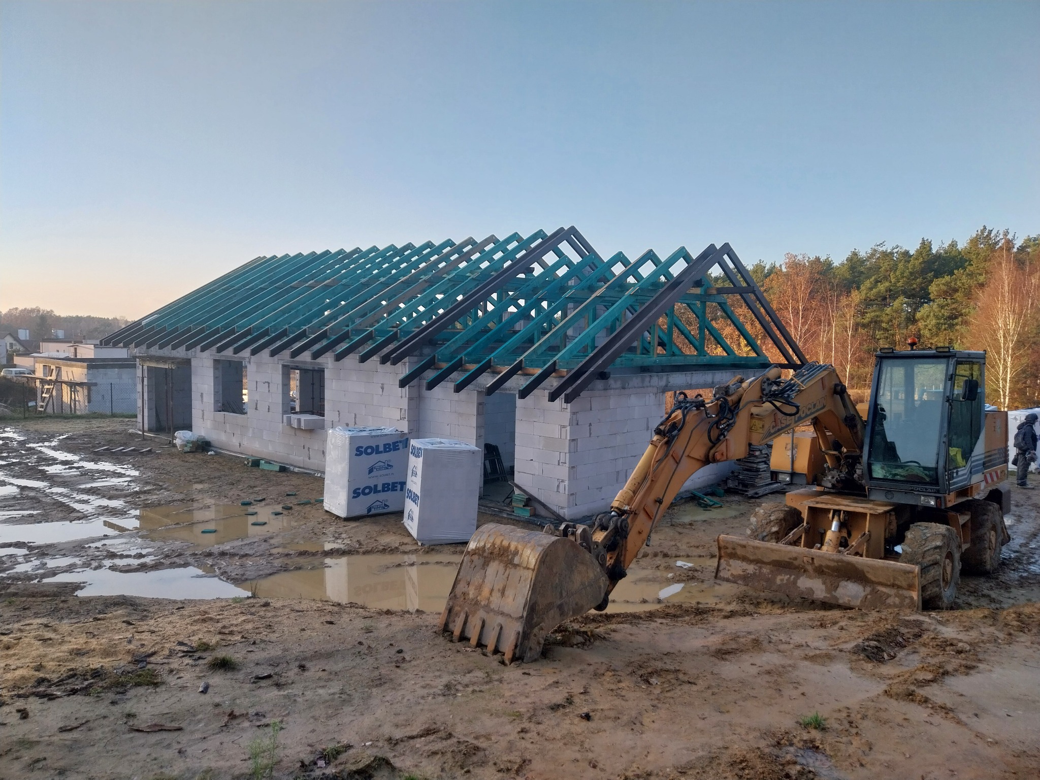 Stan surowy otwarty domu z bloczków, widoczna konstrukcja dachu z impregnowanego drewna w kolorze zielonym, na pierwszym planie koparka w błotnistym terenie, w tle las.