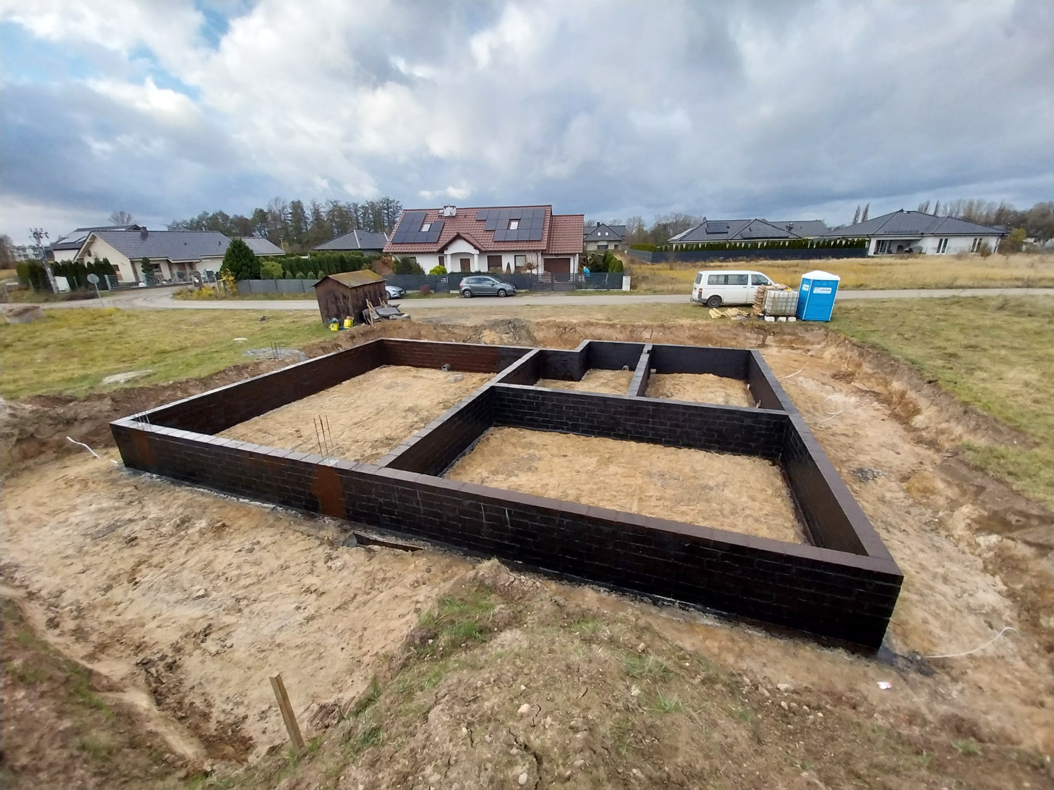 Wykop pod fundamenty domu w Ochli, z wyizolowanymi ścianami fundamentowymi z bloczków, widok z góry na tle zabudowy jednorodzinnej i pochmurnego nieba.