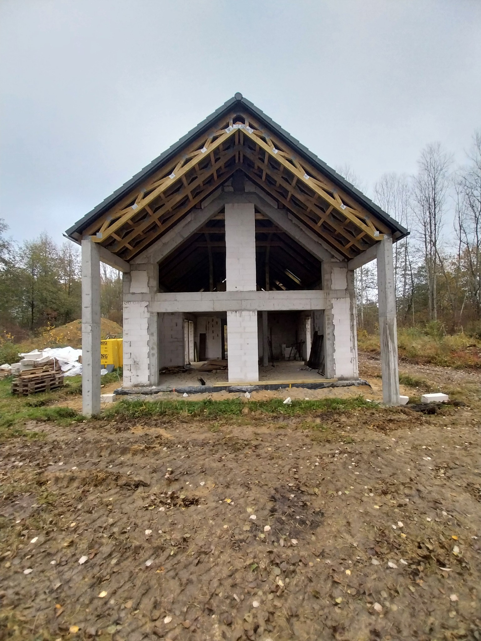 Surowy stan dwukondygnacyjnego domu z betonu komórkowego z drewnianą konstrukcją dachu, widoczne palety z materiałami budowlanymi i błotniste podłoże.