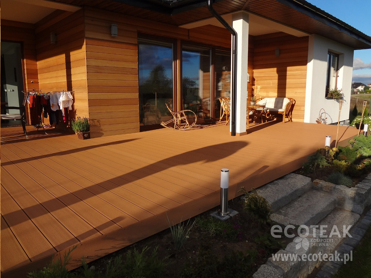 Nowoczesny taras z desek w kolorze ciepłego brązu, z widocznym drewnianym wykończeniem elewacji domu, elementami mebli ogrodowych i oświetleniem w postaci niskich lamp.