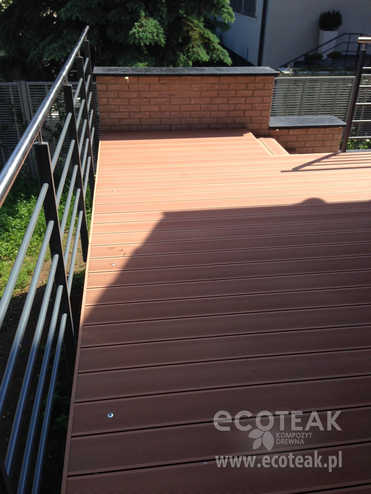 Nowoczesny taras z desek kompozytowych w odcieniach brązu, z metalową balustradą i elementami muru z cegły, wykończony z dbałością o detale.