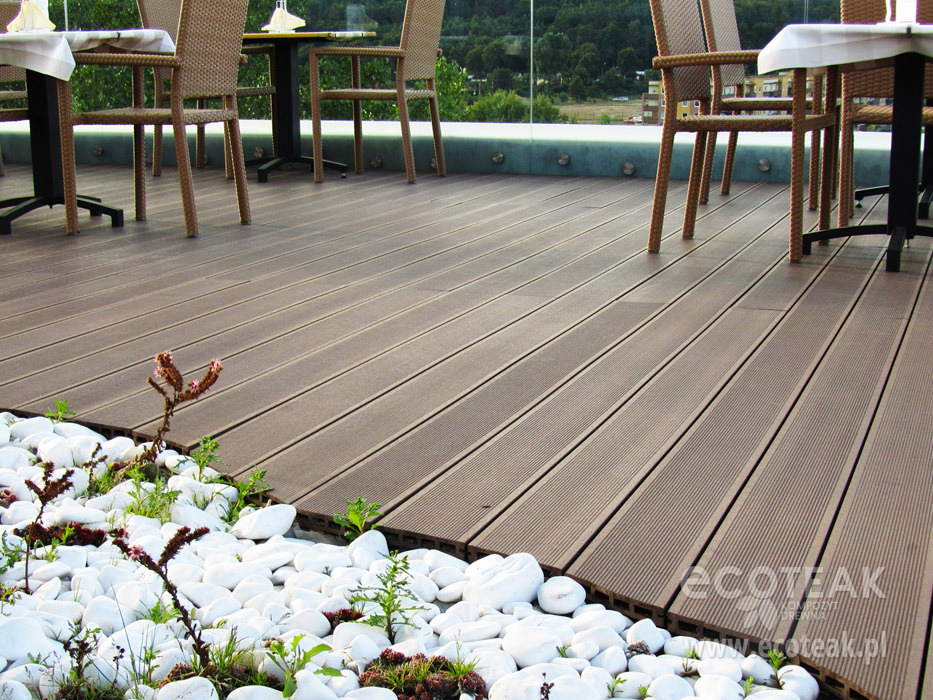 Zadaszony taras z brązowych desek, otoczony białymi kamieniami i zielenią, z widokiem na zieleń za szklaną balustradą, przygotowany do spożywania posiłków na świeżym powietrzu.