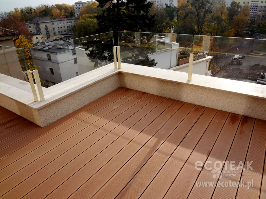 Balkon wyłożony deskami w kolorze jasnego brązu z szklaną balustradą, widok na okoliczne budynki i drzewa w jesiennych barwach, logo producenta w prawym dolnym rogu.