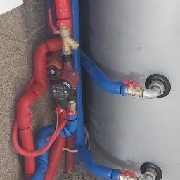 Tomasz Chabowski - Instalacja pompy ciepła z izolowanymi rurami w kolorze czerwonym i niebieskim, widoczny zawór i manometr, na tle szarego zbiornika i wzorzystej podłogi.