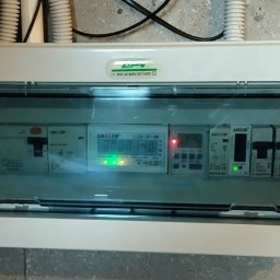 Tomasz Chabowski - Biała skrzynka rozdzielcza z przezroczystym wiekiem, zawierająca liczniki i wyłączniki, zamontowana na szarej ścianie. Widoczne kable w peszlach w górnej części.