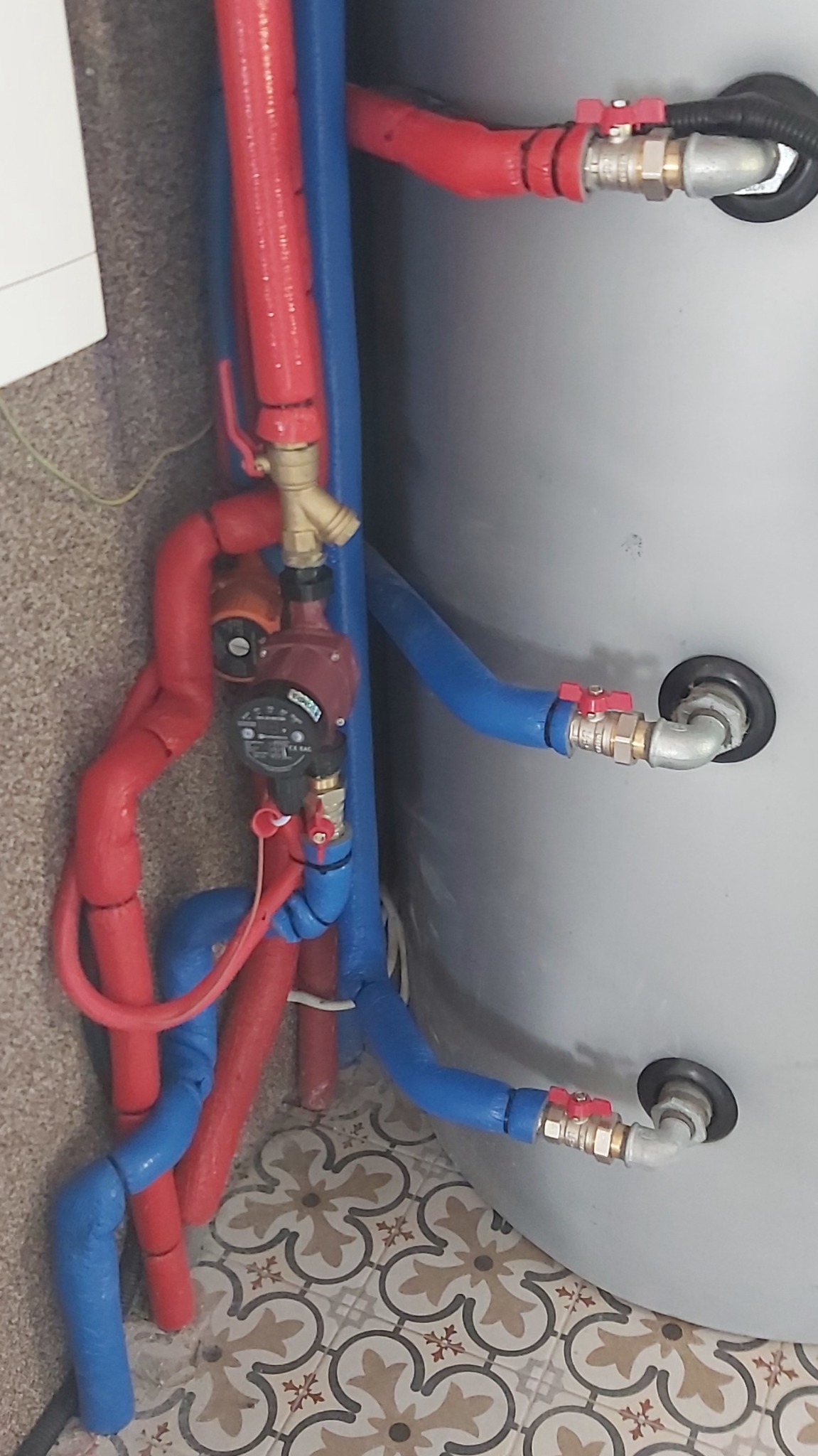 Instalacja pompy ciepła z izolowanymi rurami w kolorze czerwonym i niebieskim, widoczny zawór i manometr, na tle szarego zbiornika i wzorzystej podłogi.