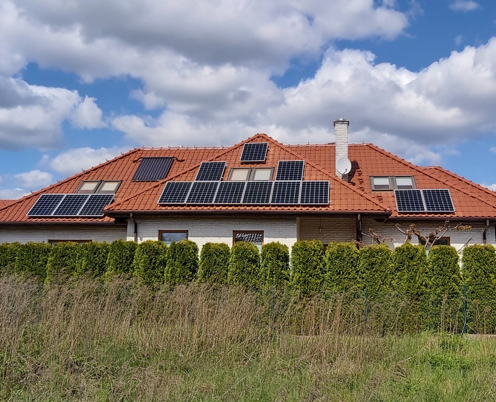 Dom jednorodzinny z czerwoną dachówką, na którym zamontowano panele fotowoltaiczne i kolektor słoneczny, widoczne okna dachowe i antena satelitarna, a w tle błękitne niebo z chmurami.