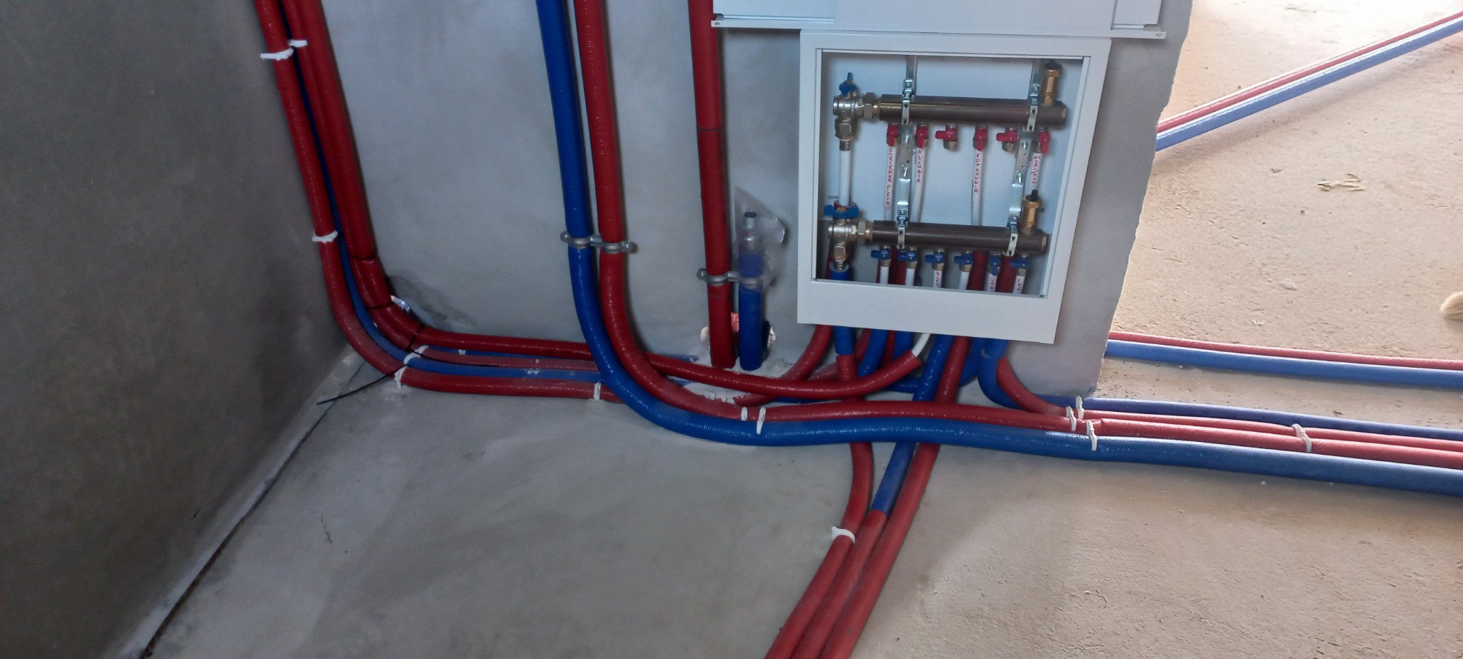 Rozdzielacz centralnego ogrzewania w białej obudowie, z licznymi zaworami i rurami PEX w kolorach czerwonym i niebieskim, przymocowanymi do ściany i podłogi w surowym stanie.