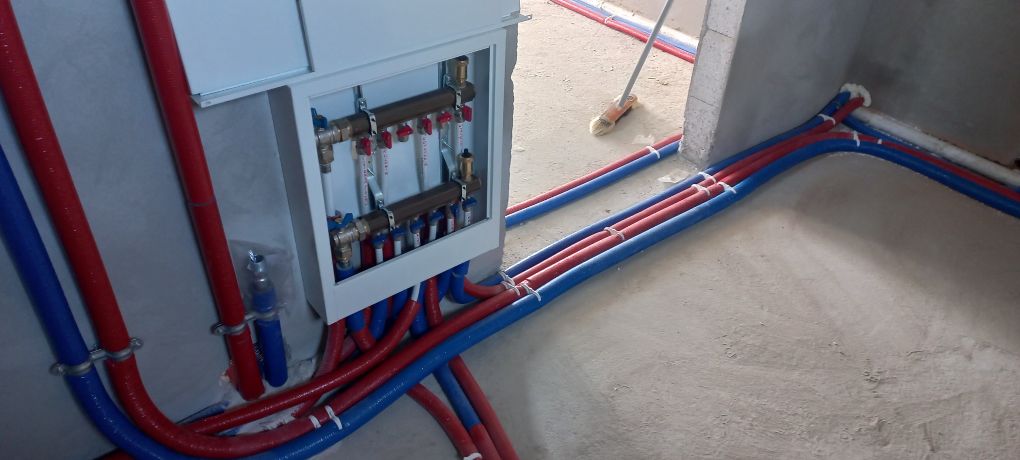 Rozdzielacz centralnego ogrzewania w obudowie, z licznymi rurami w izolacji czerwonej i niebieskiej, zamocowanymi do ściany klamrami, widoczne także fragmenty podłogi i ściany w surowym stanie.