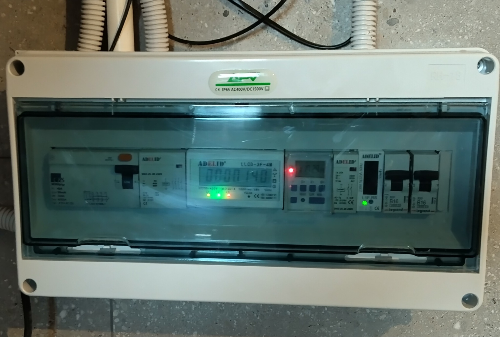 Biała skrzynka rozdzielcza z przezroczystym wiekiem, zawierająca liczniki i wyłączniki, zamontowana na szarej ścianie. Widoczne kable w peszlach w górnej części.