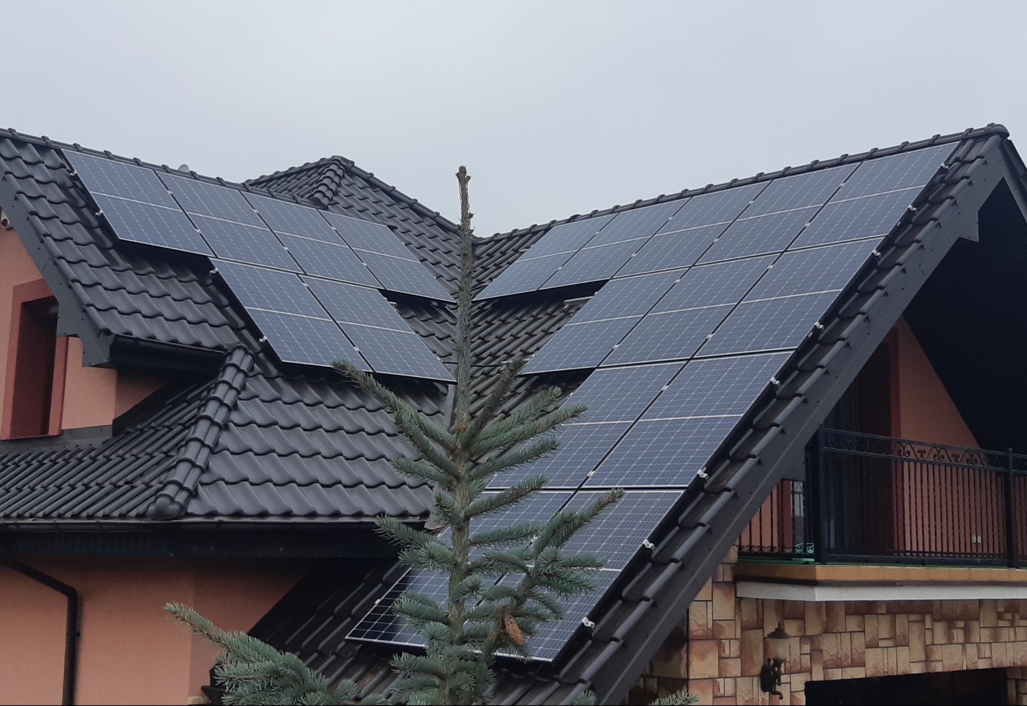 Panele fotowoltaiczne zamontowane na dachu domu jednorodzinnego z czarną dachówką, widoczne zza świerku.