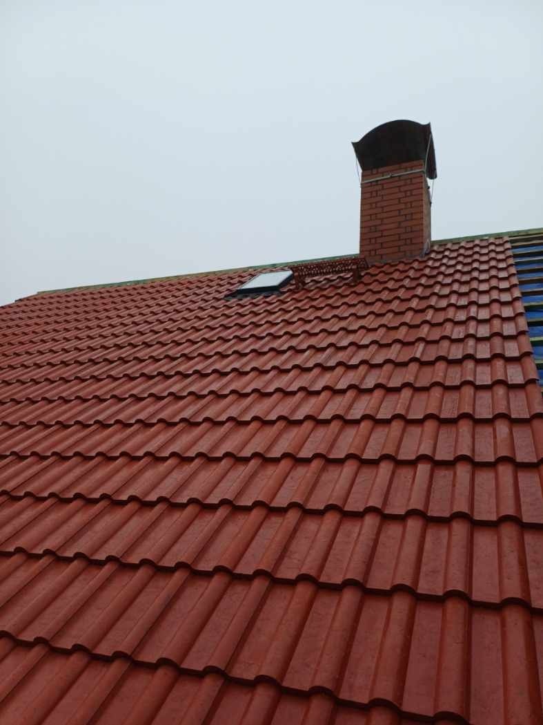 Czerwony dach z ceramicznych elementów z kominem i oknem dachowym, fragment nieukończonego pokrycia z widoczną membraną.