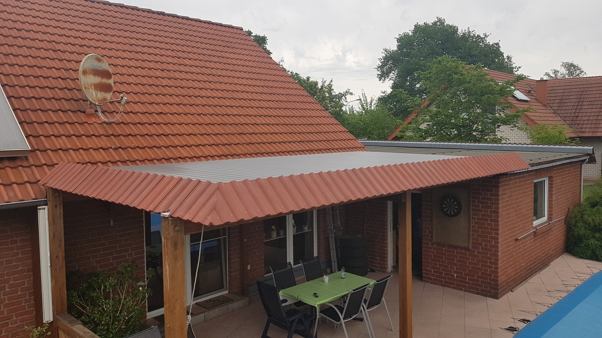 Zadaszona, ceglana weranda z czerwonym, falistym pokryciem dachowym, widok z góry; w tle dom z dachem pokrytym dachówką ceramiczną i zardzewiałą anteną satelitarną.