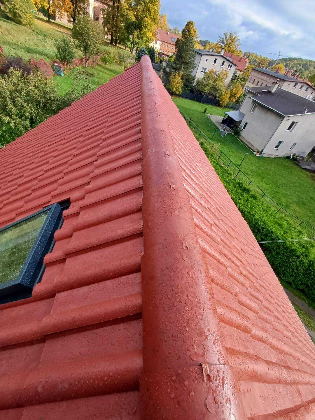 Czerwony dach z ceramiczną dachówką, widoczne okno dachowe i krople wody po deszczu, w tle zabudowa jednorodzinna i zieleń.
